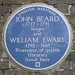 A Blue plaque on a brick wall with the words "John Beard C1717&nbsp;– 1791 Singer and William Ewart 1798&nbsp;– 1861 Promoter of Public Libraries