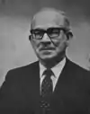 A photographic portrait of a man wearing glasses and a suit