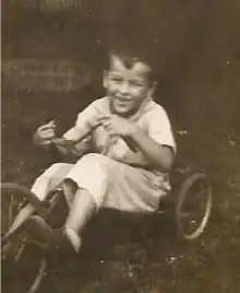 Toddler Ted Stevens rides on a tricycle. Stevens has a smile. The image is in sepiatone.